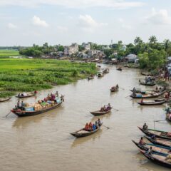 Barcos tradicionais navegando nos rios do delta em Bangladesh com campos agrícolas ao fundo sob luz natural.